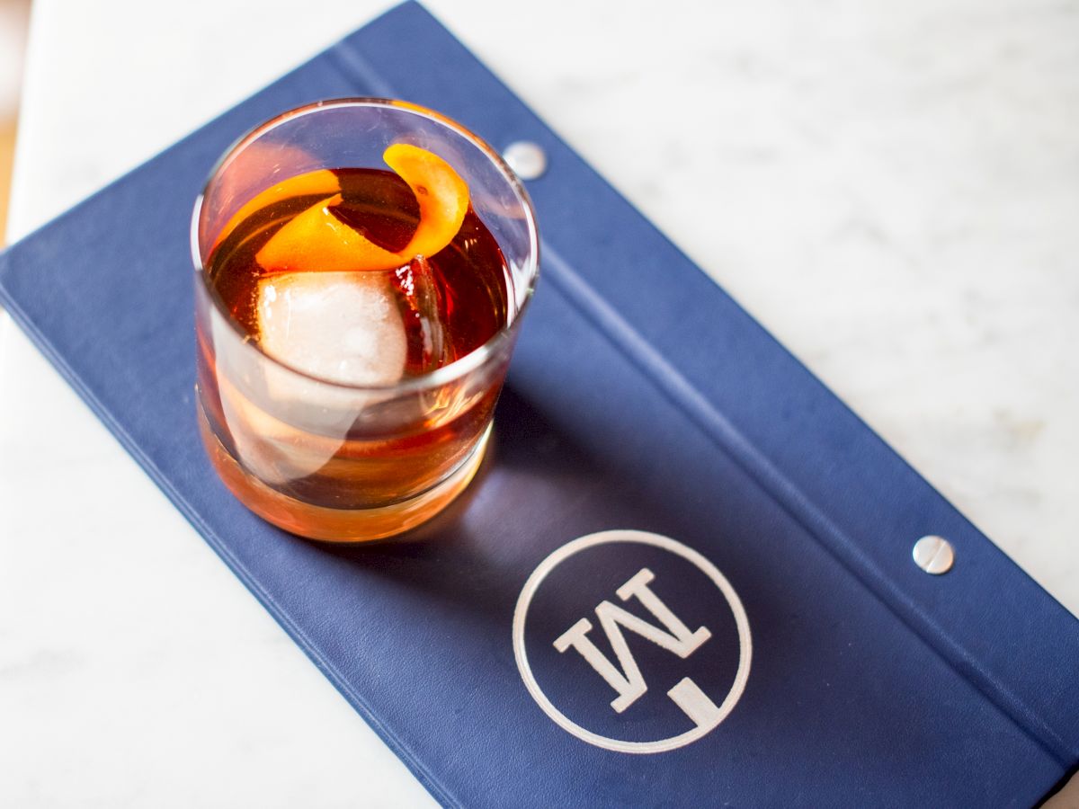 A glass of amber whiskey with a lemon twist sits on a blue menu or notebook branded with a white circular logo, atop a white marble surface.