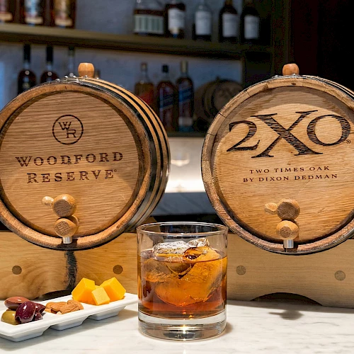 Two wooden whiskey barrels labeled Woodford Reserve and 2XO, with a glass of whiskey and a plate of small appetizers on a bar.