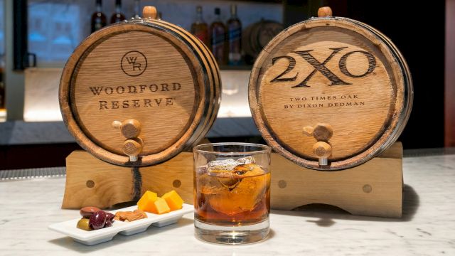 Two wooden whiskey barrels labeled Woodford Reserve and 2XO, with a glass of whiskey and a plate of small appetizers on a bar.