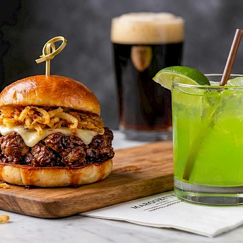 A juicy double-stack burger with melted cheese and crispy onions on a wooden board, beside a lime-green cocktail on the table, delicious.