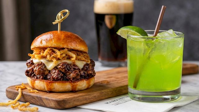 A juicy double-stack burger with melted cheese and crispy onions on a wooden board, beside a lime-green cocktail on the table, delicious.