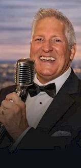 A smiling man in a tuxedo sings into a vintage microphone, holding it with one hand against a cityscape backdrop.