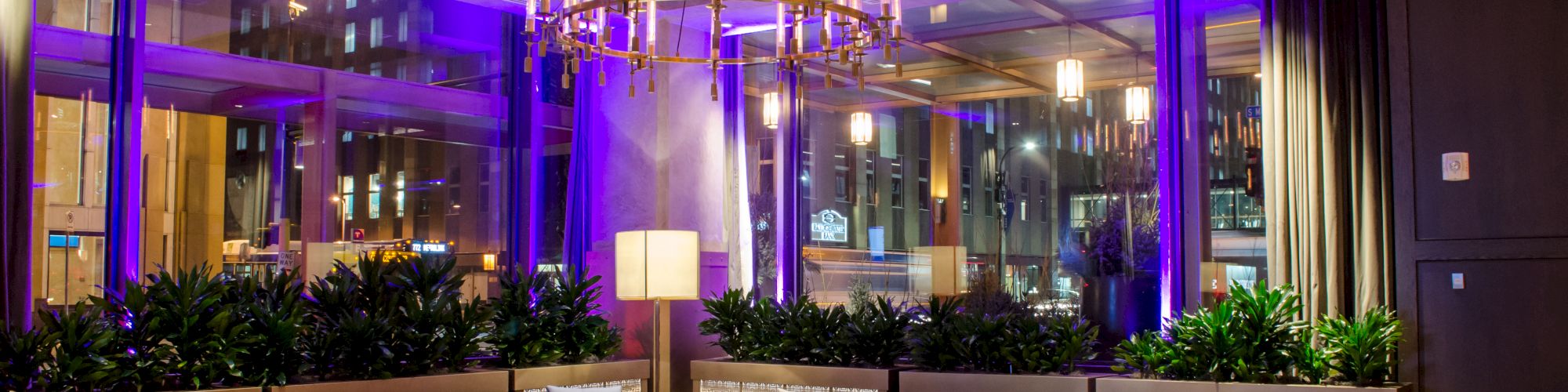 A modern hotel lobby with purple lighting, a chandelier, modular gray sofas, glass coffee tables, greenery, and large windows.