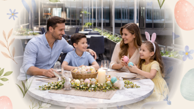 A family scene: parents with two children enjoy a sunny outdoor Easter meal at a round table, decorated with eggs and flowers, smiling together.