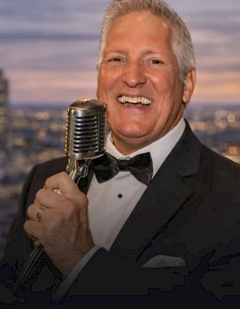 A smiling man in a tuxedo sings into a vintage microphone, holding it with one hand against a cityscape backdrop.