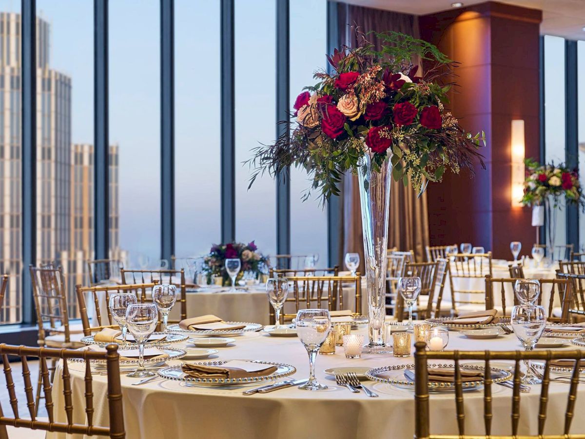 A elegant banquet setup in a high-rise venue, round tables with white linens, tall floral centerpieces, and neatly arranged place settings, overlooking a cityscape.