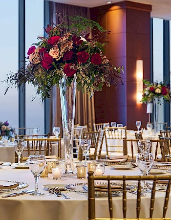 A elegant banquet setup in a high-rise venue with round tables, gold chairs, white linens, glassware, and tall floral centerpieces, overlooking a city skyline.