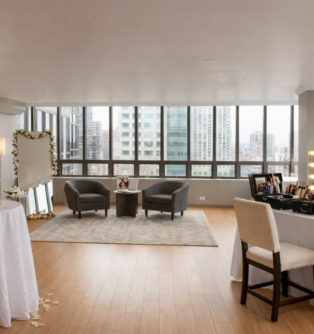 A bright, stylish makeup room with two vanity tables, mirror lights, chairs, and seating area by large windows overlooking the city.