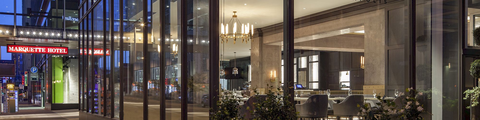 A modern glass hotel lobby at night with &ldquo;Marquette&rdquo; signage, warm lighting, and visible seating inside; outdoor sidewalk and red neon signs nearby.