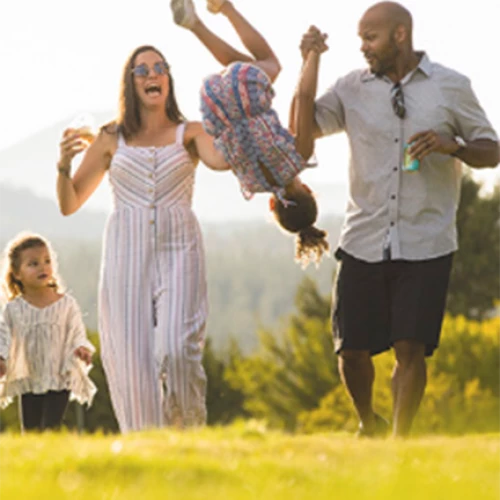 A family of four is outdoors on a sunny day. The parents are swinging one child by the arms while the other child walks beside them, smiling.