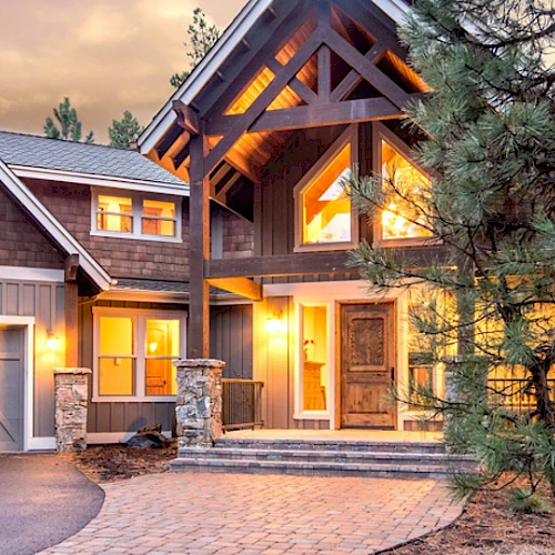 A large, well-lit house with a modern rustic design, large windows, a two-car garage, and surrounded by trees at dusk completes this image.