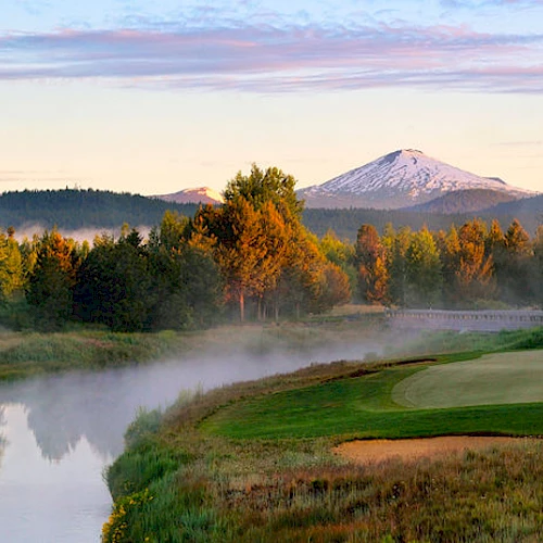 A scenic landscape with a golf course near a river and trees, mountains in the background, and a clear sky with light clouds at sunrise or sunset.