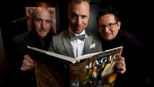 Three men in dark suits and a bow tie sit together, smiling while reading an open comic/magazine under warm lamp light.