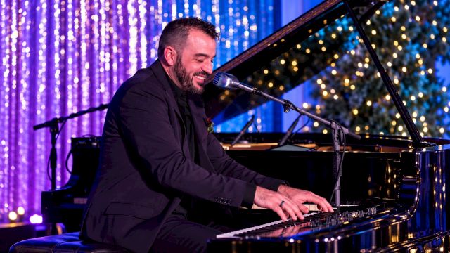 A man in a suit plays a grand piano on stage, smiling into a microphone under colorful lights.