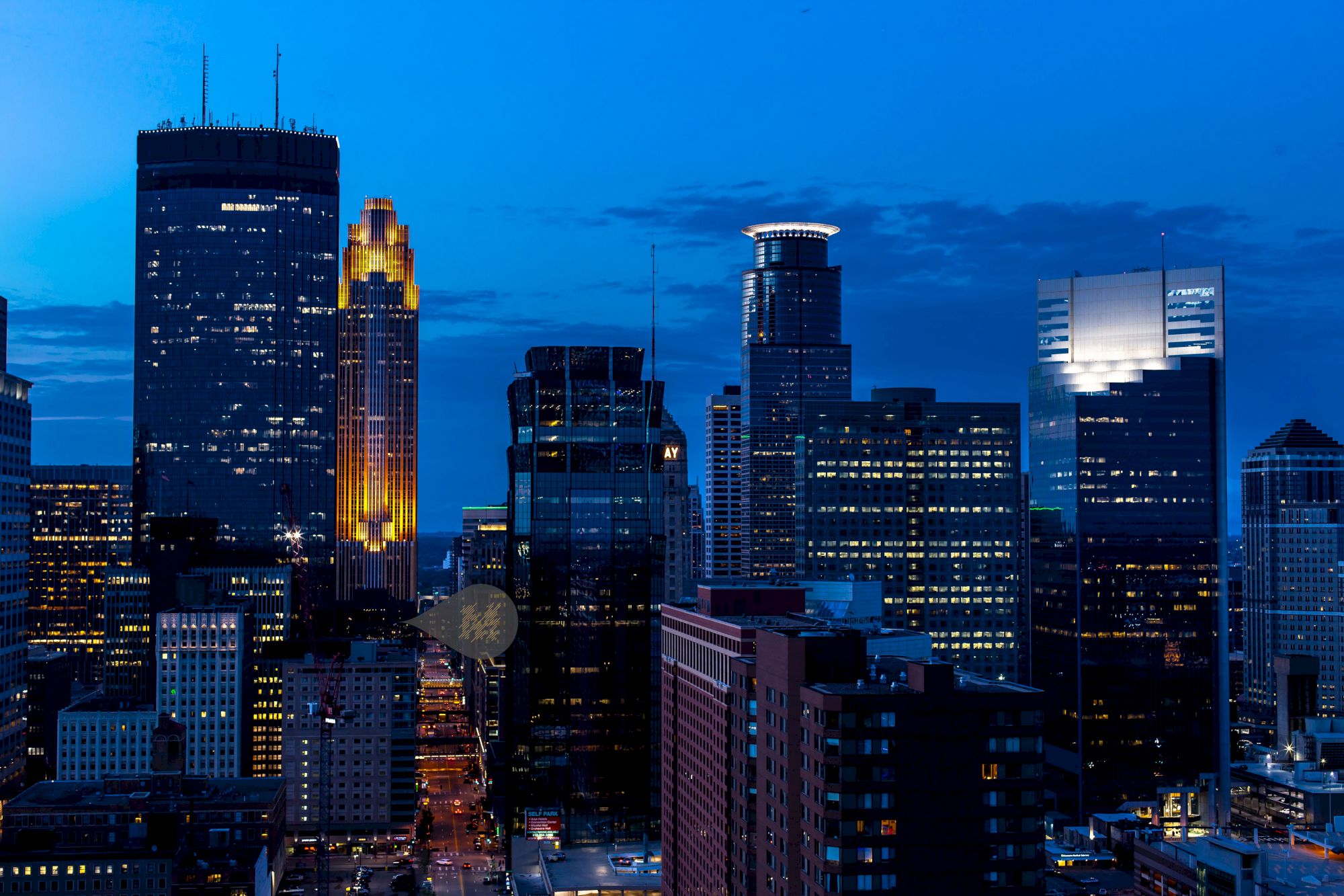 A vibrant city skyline at dusk with illuminated skyscrapers and a deep blue sky in the background, creating a striking urban night scene.