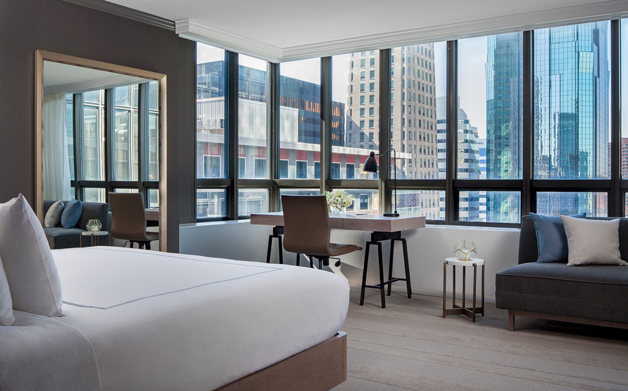 A modern bedroom with a large bed, desk, and lounge area with cityscape views through large windows, featuring sleek furniture and neutral tones.