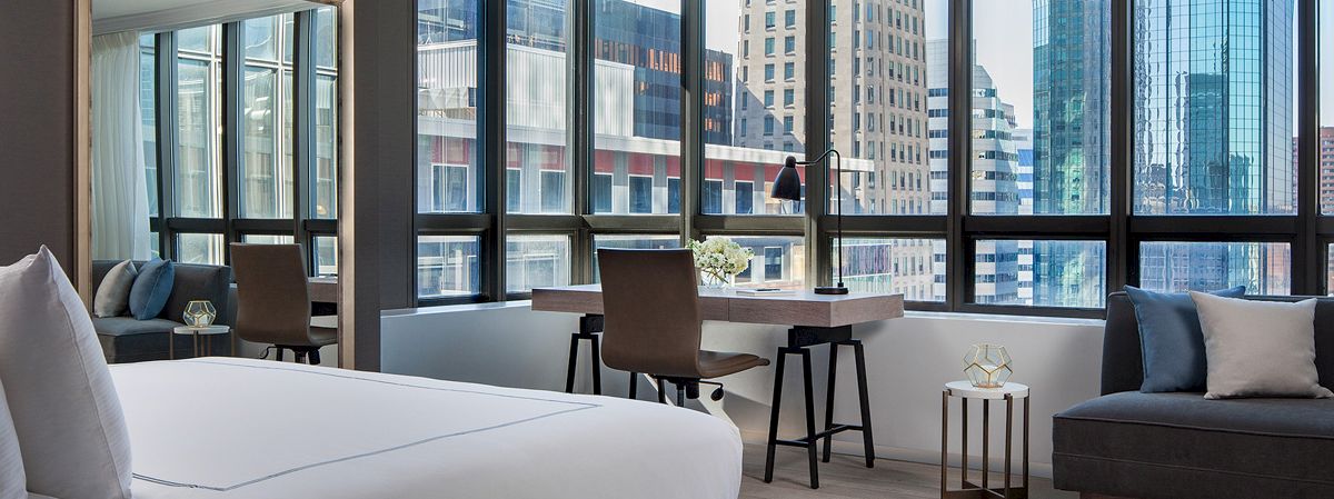 A modern bedroom with a large bed, desk, and lounge area with cityscape views through large windows, featuring sleek furniture and neutral tones.
