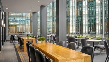 The image shows a modern indoor seating area with wooden tables, black chairs, and large windows letting in natural light.