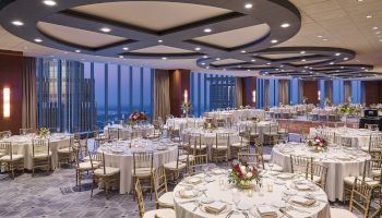 A large, elegant banquet hall with round tables set for an event, featuring floor-to-ceiling windows offering a city view and modern ceiling design.