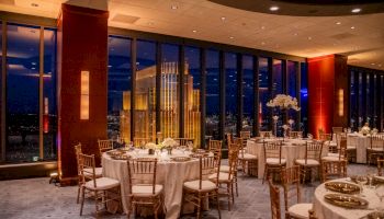 An elegant event space with round tables, chairs, and a city view through tall windows at night.