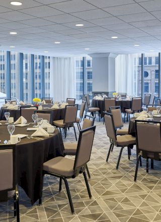 A banquet room with round tables set up for an event, featuring tablecloths, chairs, glassware, and folded napkins, with city views through large windows.