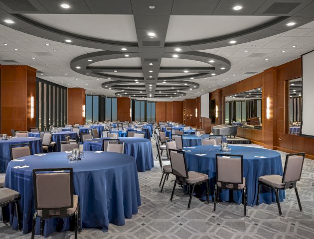 A conference room set up with round tables covered in blue tablecloths and surrounded by chairs, with modern lighting and decor, and a large screen.