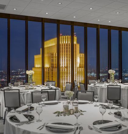 A luxurious banquet room with elegantly set tables overlooks a cityscape highlighted by a brightly lit, golden building at night through large windows.