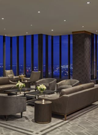 A modern, luxurious lounge with sofas and chairs, large windows showcasing a night cityscape, and a sleek, patterned carpet is shown in the image.