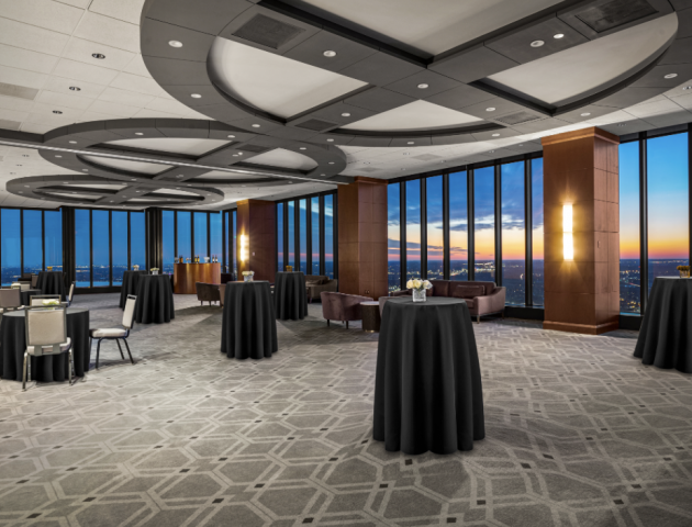 A spacious, elegantly designed event room with round tables covered in black cloths, surrounded by large windows displaying a sunset view.