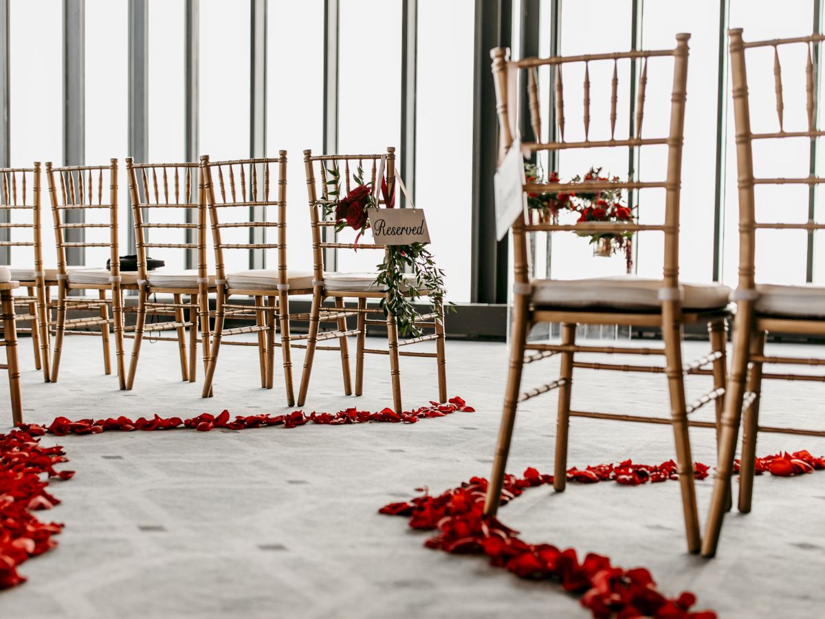 The image shows a wedding ceremony setup with wooden chairs and a curved path lined with red rose petals. A chair has a sign with 