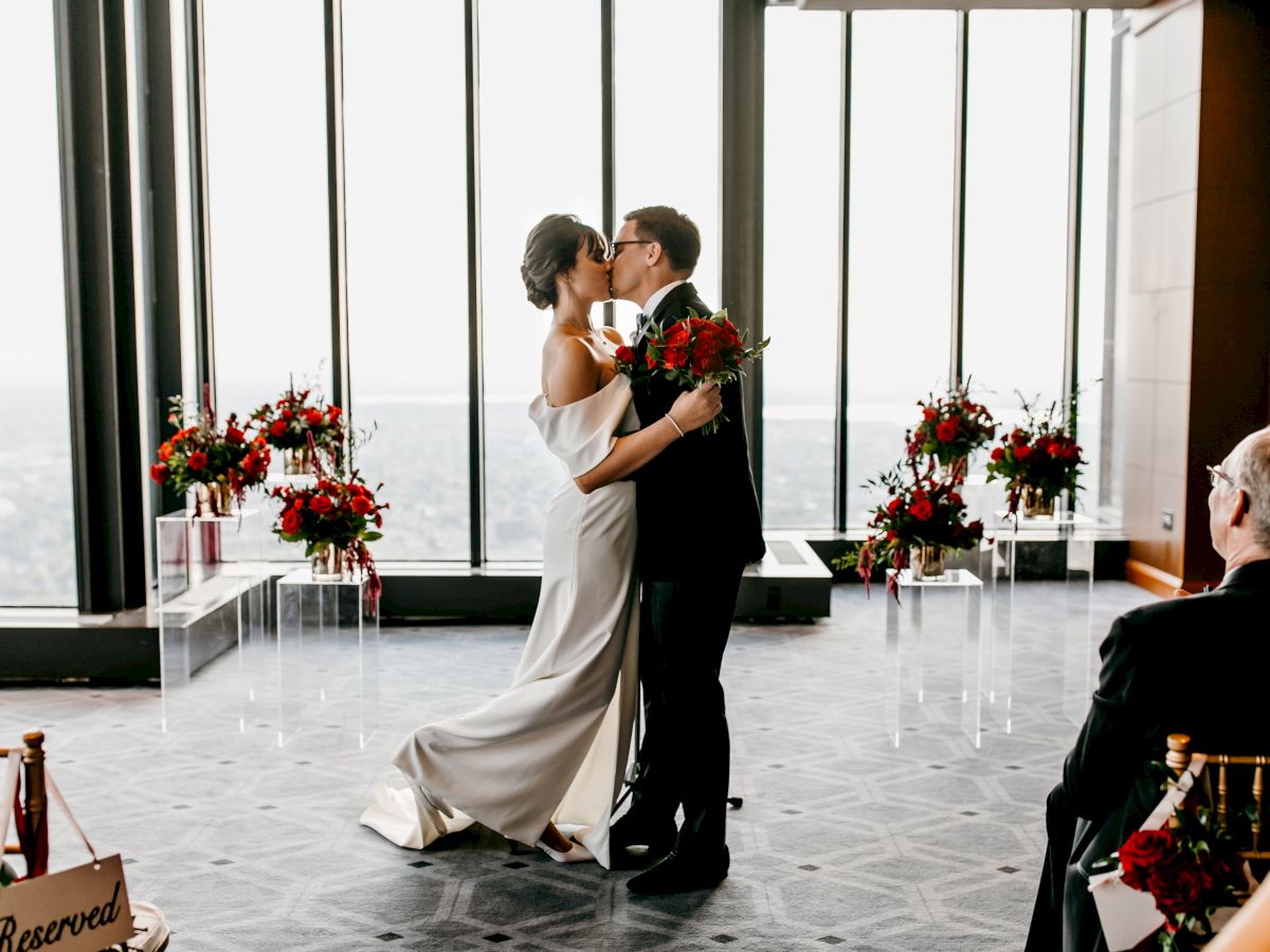 A couple is sharing a kiss during a wedding ceremony in a modern venue with large windows and floral decorations.