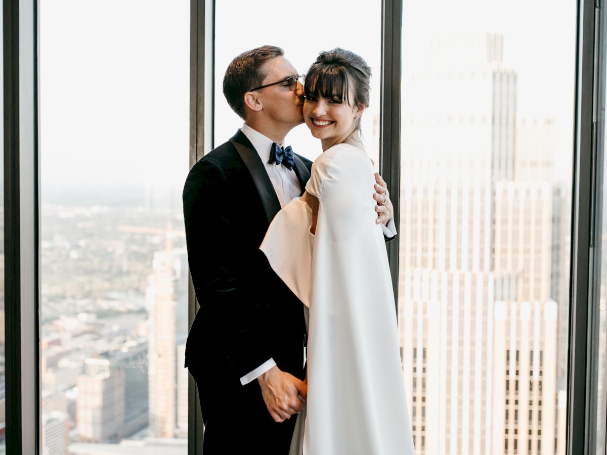 A couple dressed in formal wedding attire embrace and smile while standing in front of a cityscape view from a high-rise building's windows.