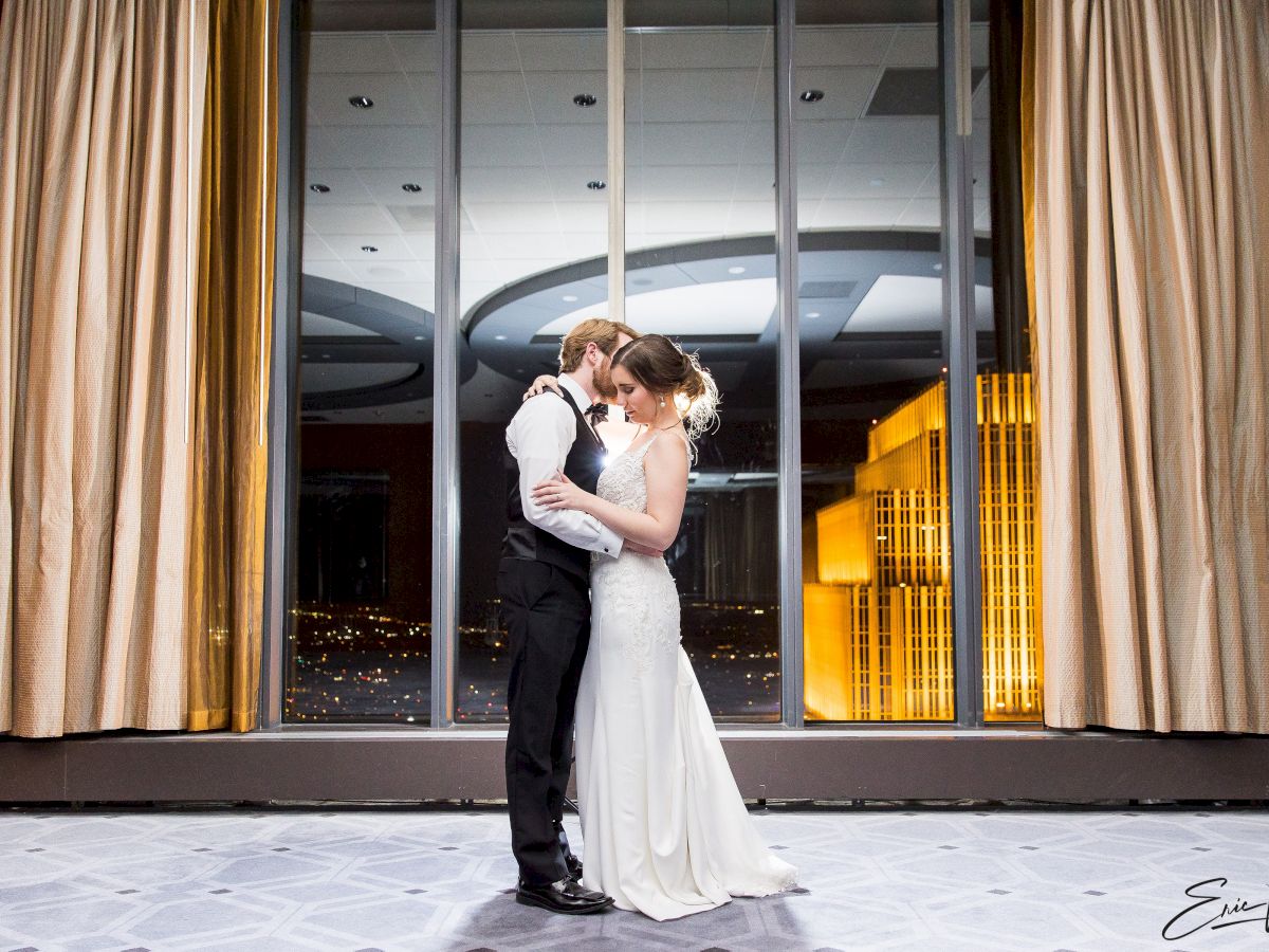 A couple in formal attire embracing in front of large windows with city lights in the background, framed by curtains, in an elegant setting.