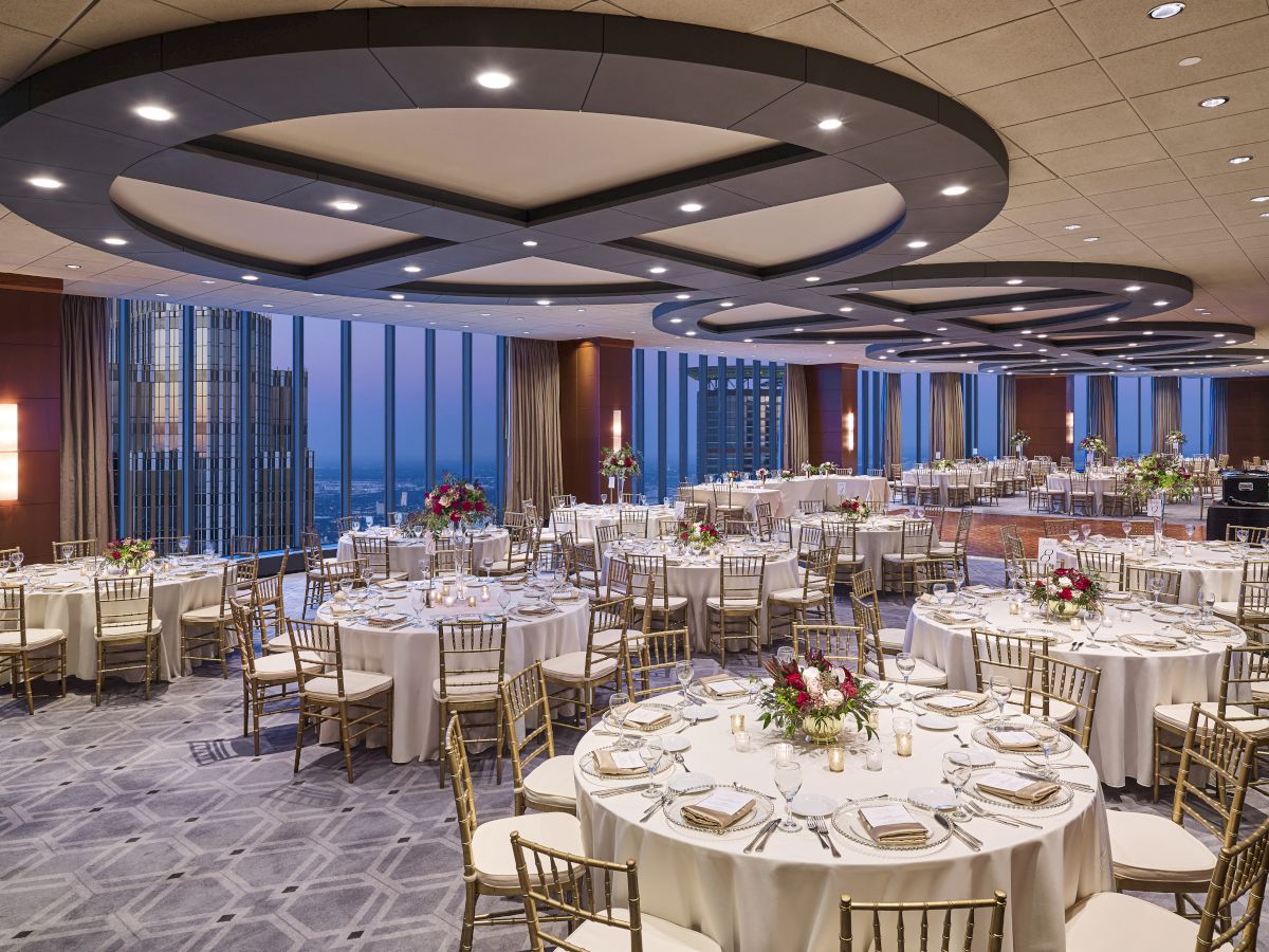 A spacious, elegant banquet hall setup with round tables, chairs, floral centerpieces, and large windows overlooking a cityscape during the evening.