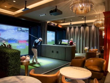 A person is playing golf in an indoor simulator. The room is stylishly furnished with chairs and lighting, creating an upscale atmosphere.