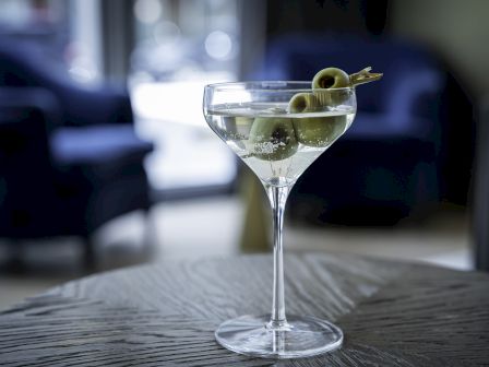 A martini glass filled with a cocktail and garnished with green olives sits on a wooden table with blurred chairs in the background.