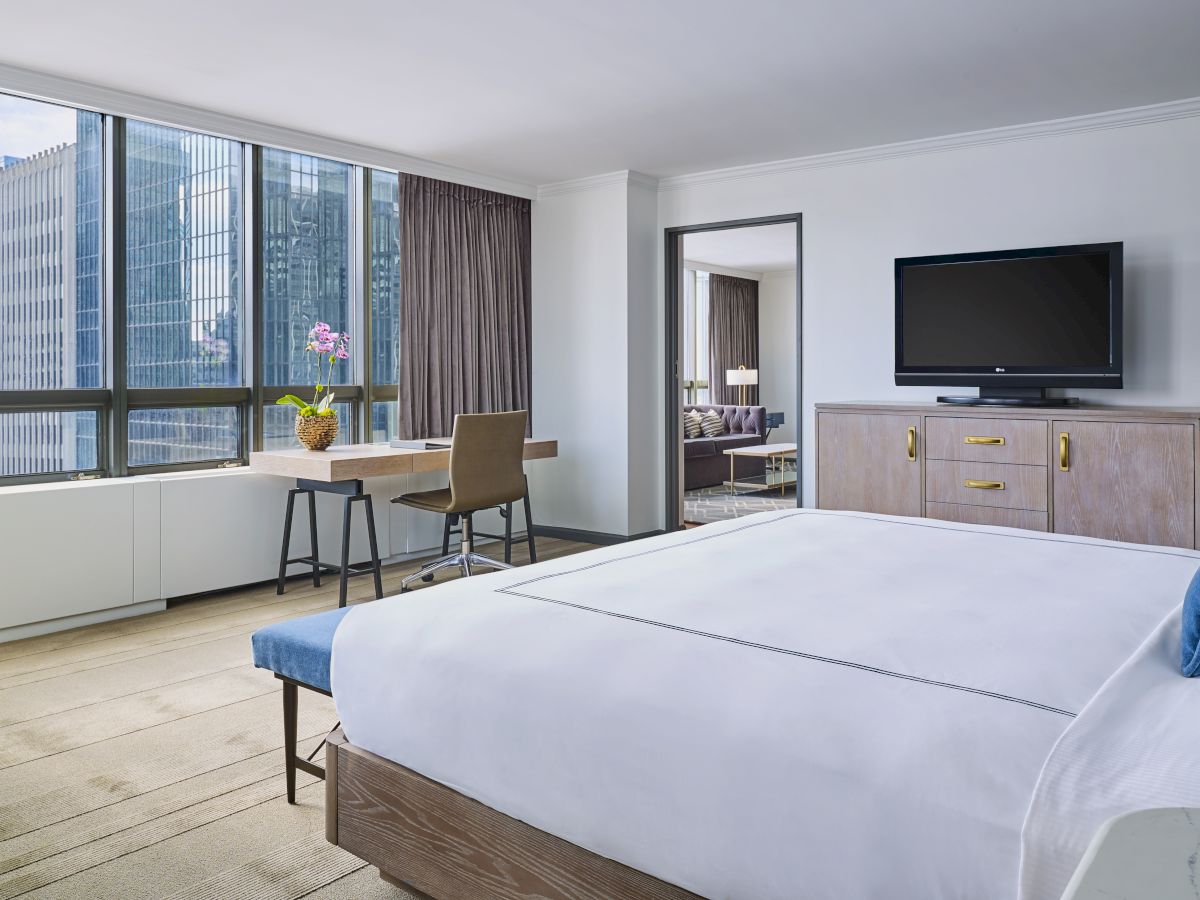 A modern hotel room with a large bed, a desk by the window, a TV on a dresser, and large windows showing a cityscape outside.