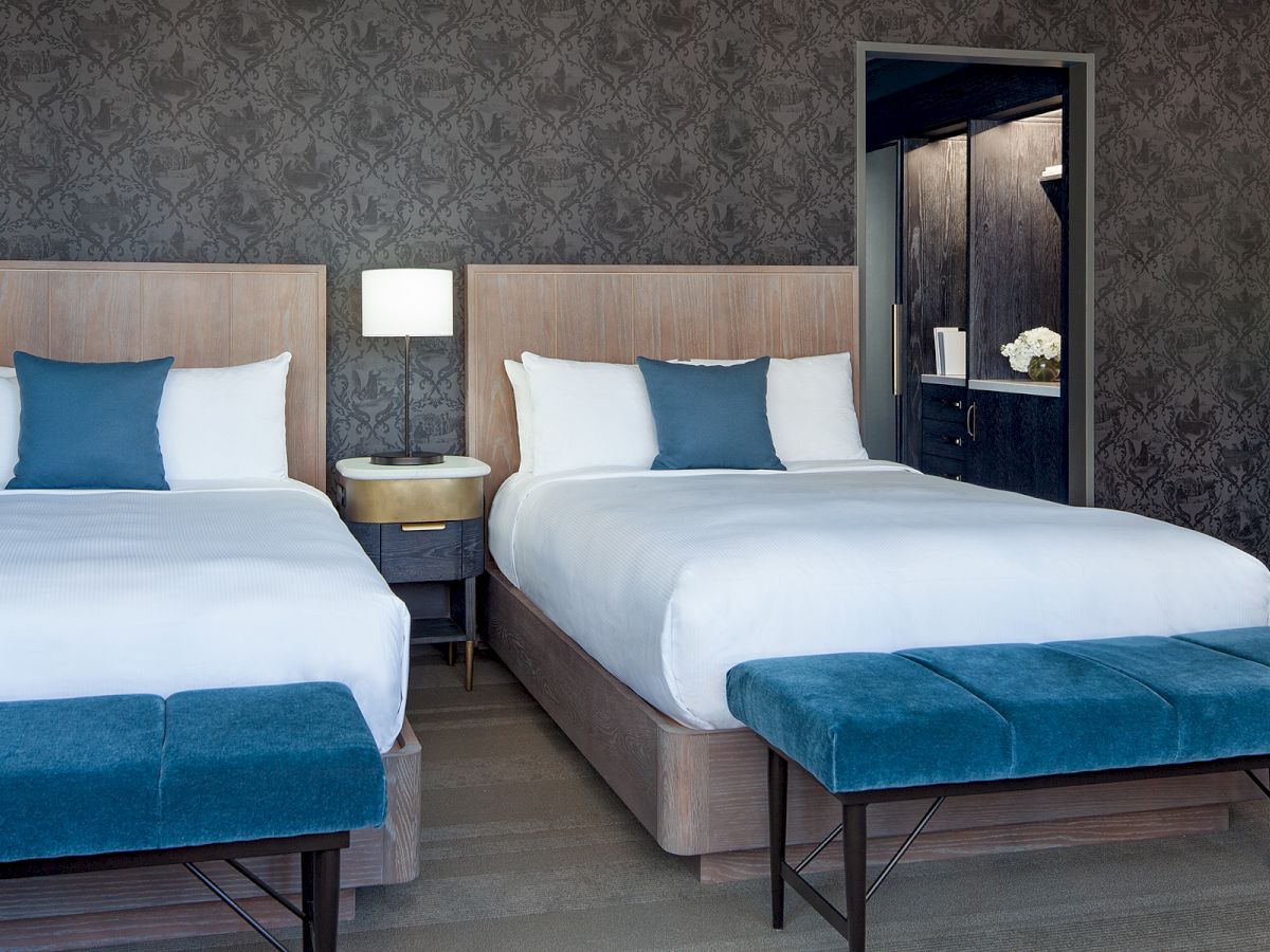 The image shows a modern hotel room with two double beds, blue accent pillows, and benches at the foot, a bedside table, and a lamp.