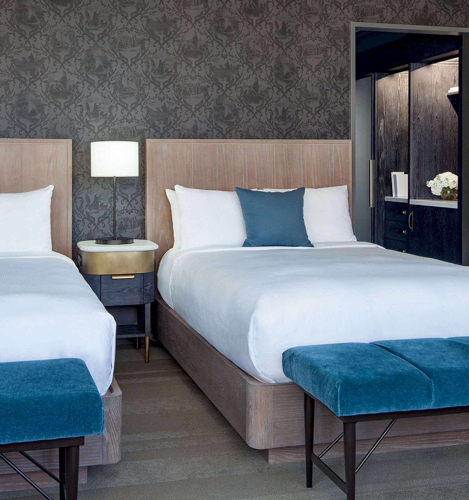 The image shows a modern hotel room with two double beds, blue accent pillows, and benches at the foot, a bedside table, and a lamp.