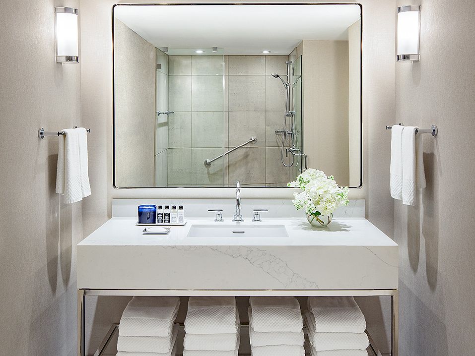 A modern bathroom features a large mirror, a sink with a flower vase, neatly folded towels below, and a walk-in shower in the background.