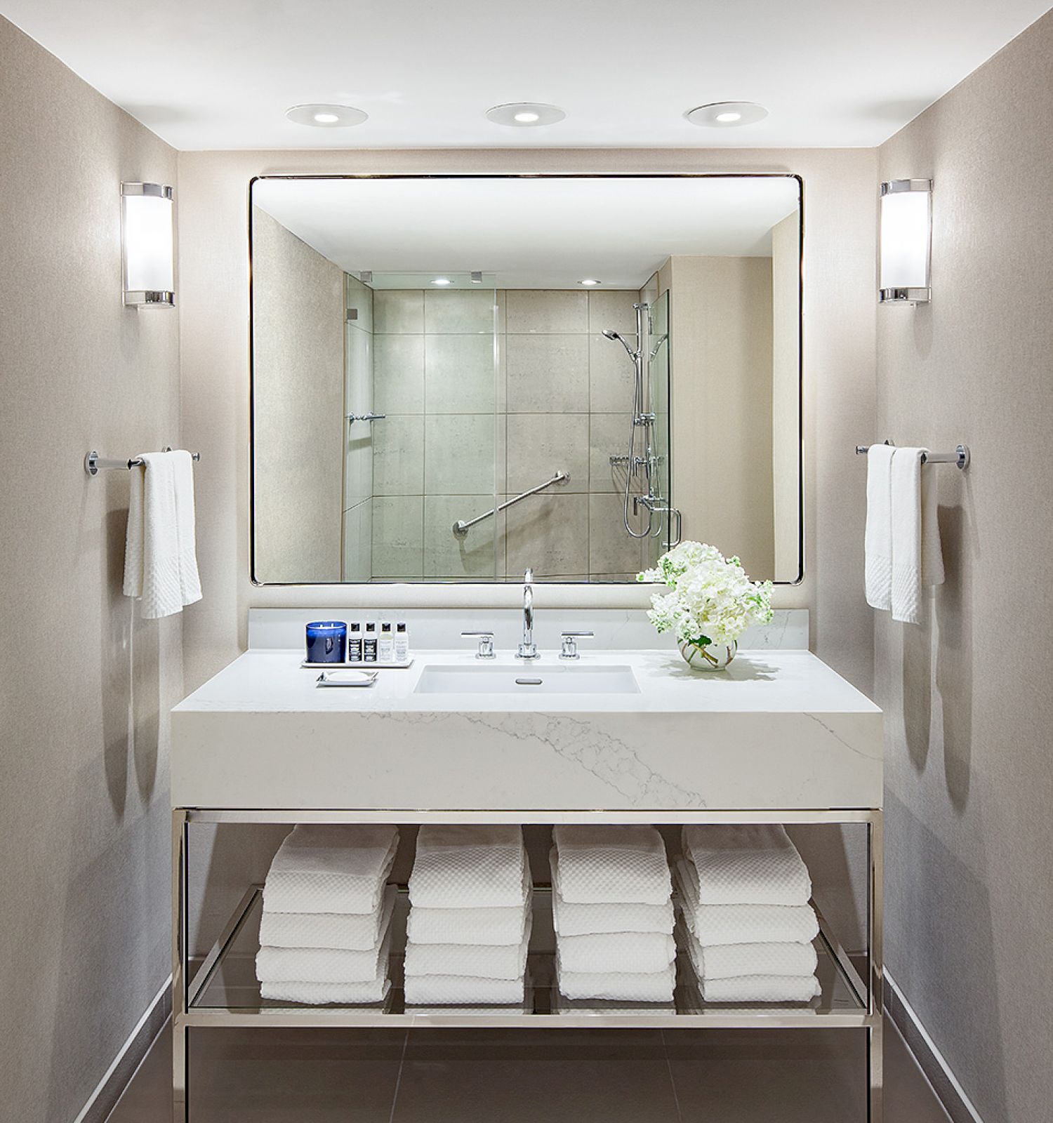 A modern bathroom features a large mirror, a sink with a flower vase, neatly folded towels below, and a walk-in shower in the background.