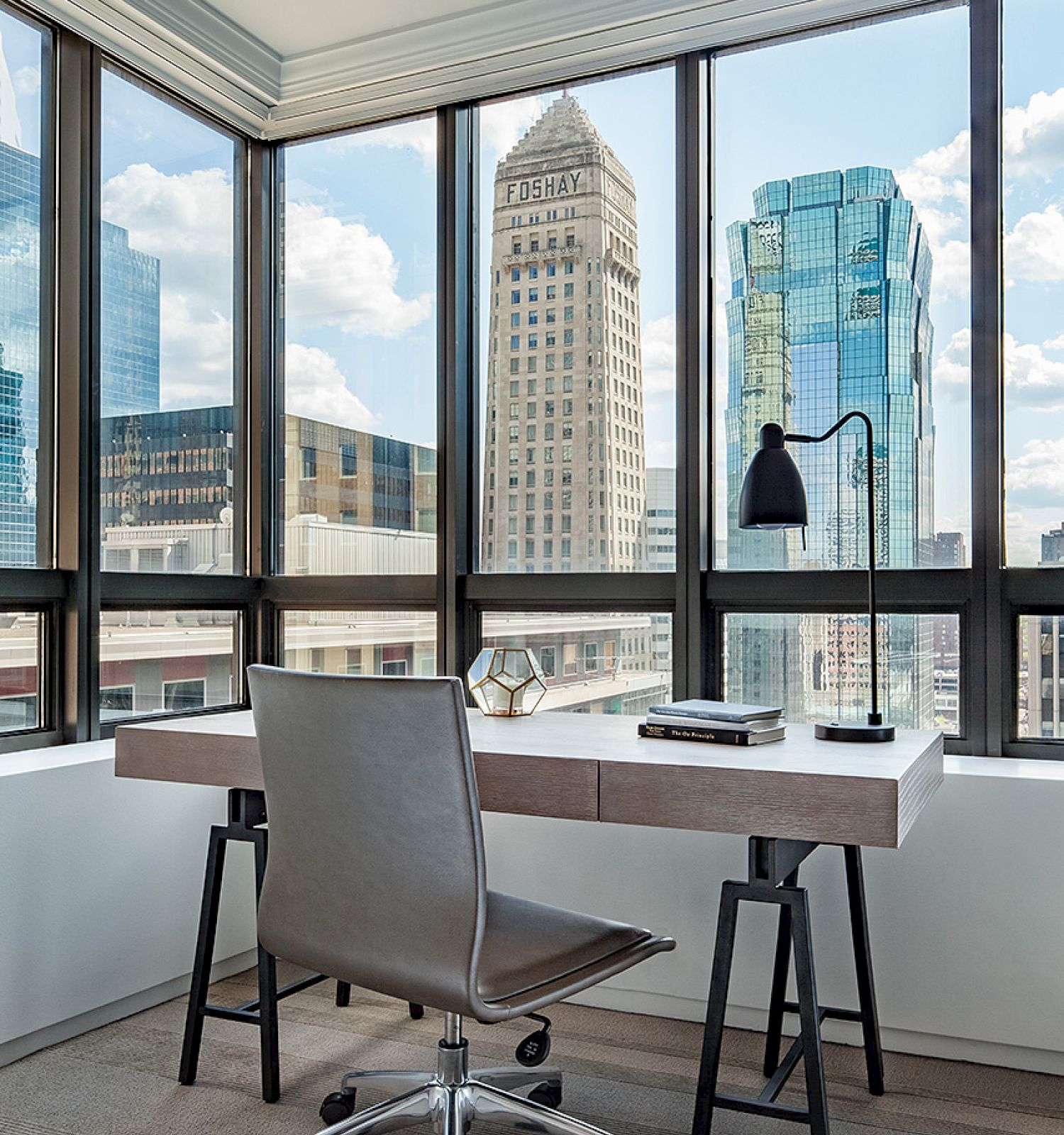 A modern office setup with a desk, chair, desk lamp, and city skyline visible through large windows in the background.