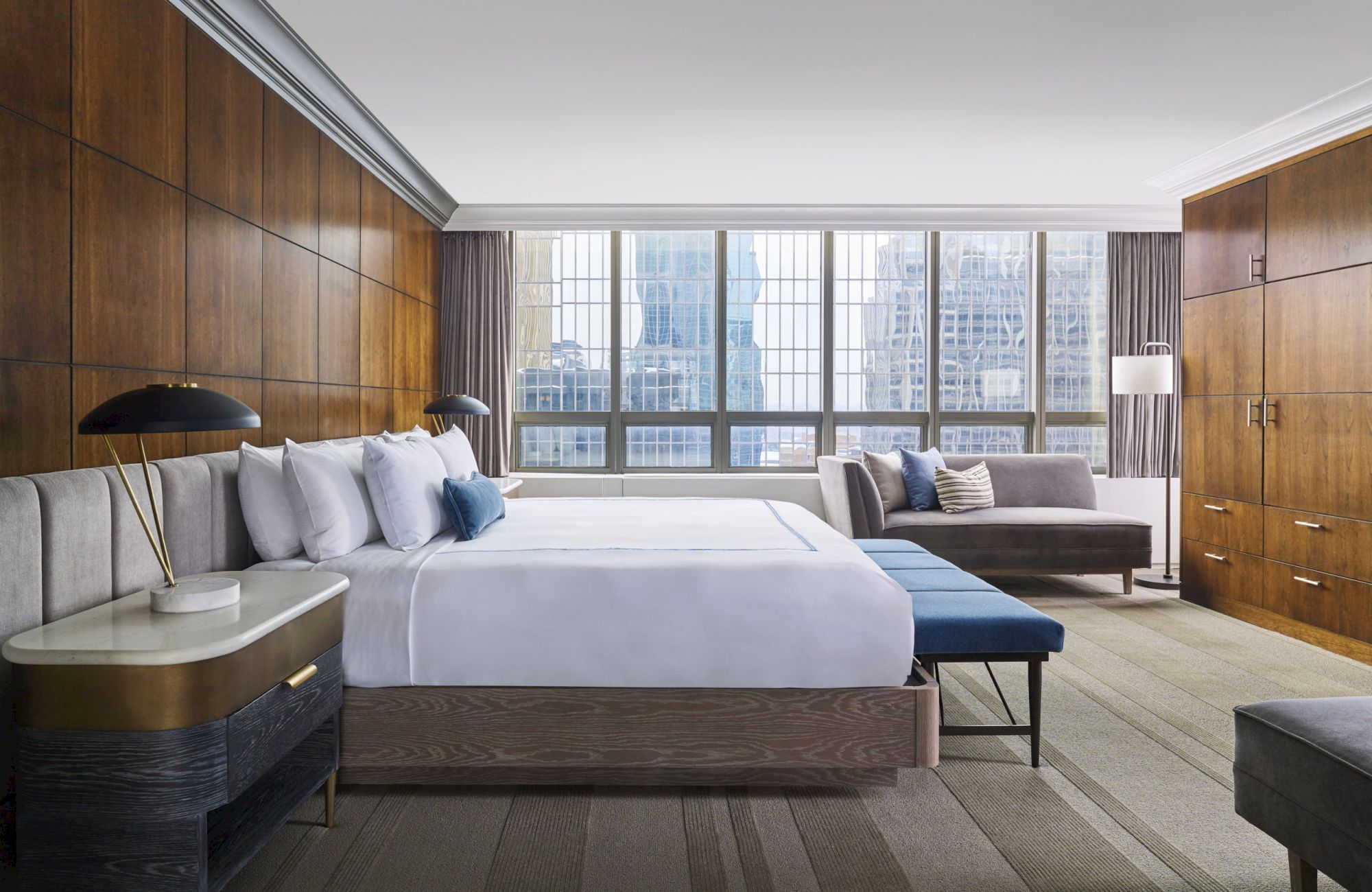 This image shows a modern hotel room with a large bed, wooden paneling, bedside tables, a sofa, a bench, and a city view through large windows.