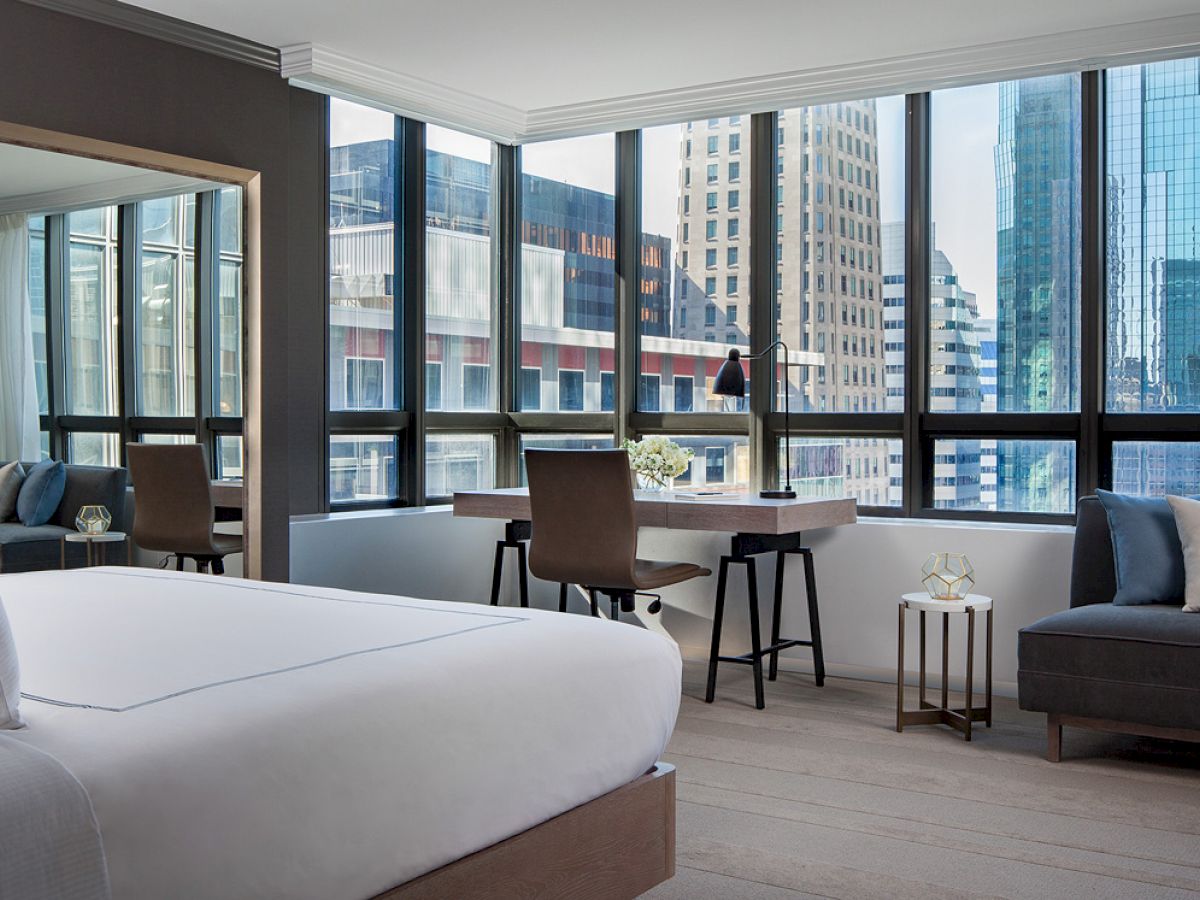 A modern hotel room with a large bed, a desk and chair by the window, a sofa, and a cityscape view through large windows.