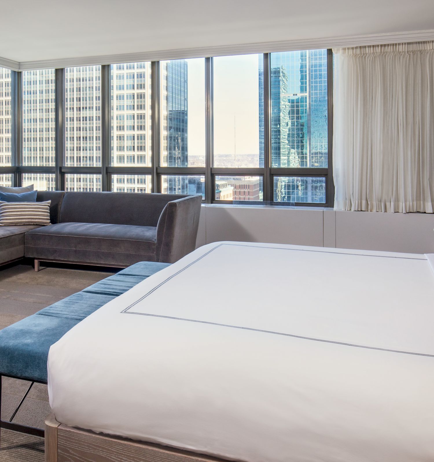 A modern hotel room with a large bed, a sectional sofa, a side table, and a city skyline view from the windows, ending the sentence.