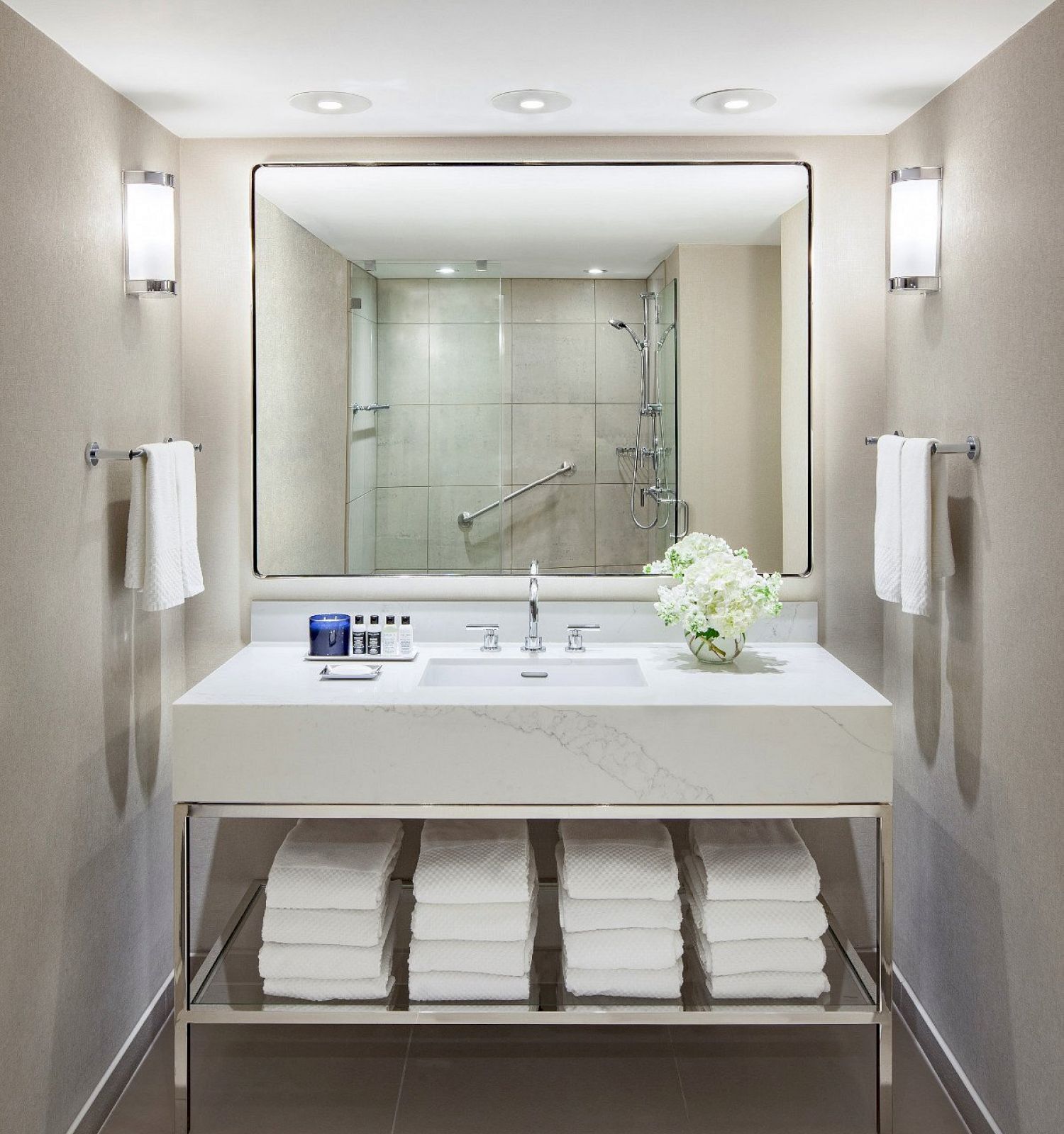 A modern bathroom with a large mirror, sink, towels beneath the counter, a flower arrangement, and a glass-enclosed shower in the background.