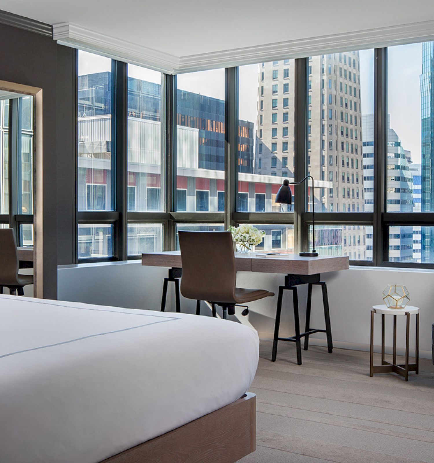 A modern hotel room features a bed, a desk, a mirror, and a seating area by large windows with a cityscape view in the background.