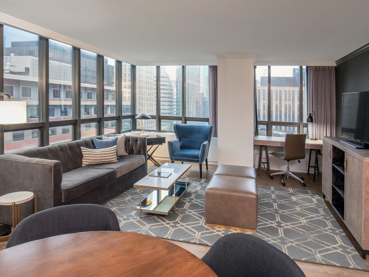 A modern living room with large windows, a couch, chairs, TV, desk, and city view. The room has a clean, elegant design and ample natural light.