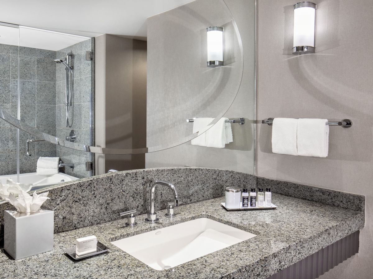 Modern bathroom with granite countertop, sink, tissue box, soap, toiletries, towels on rack, large mirror, and shower area.