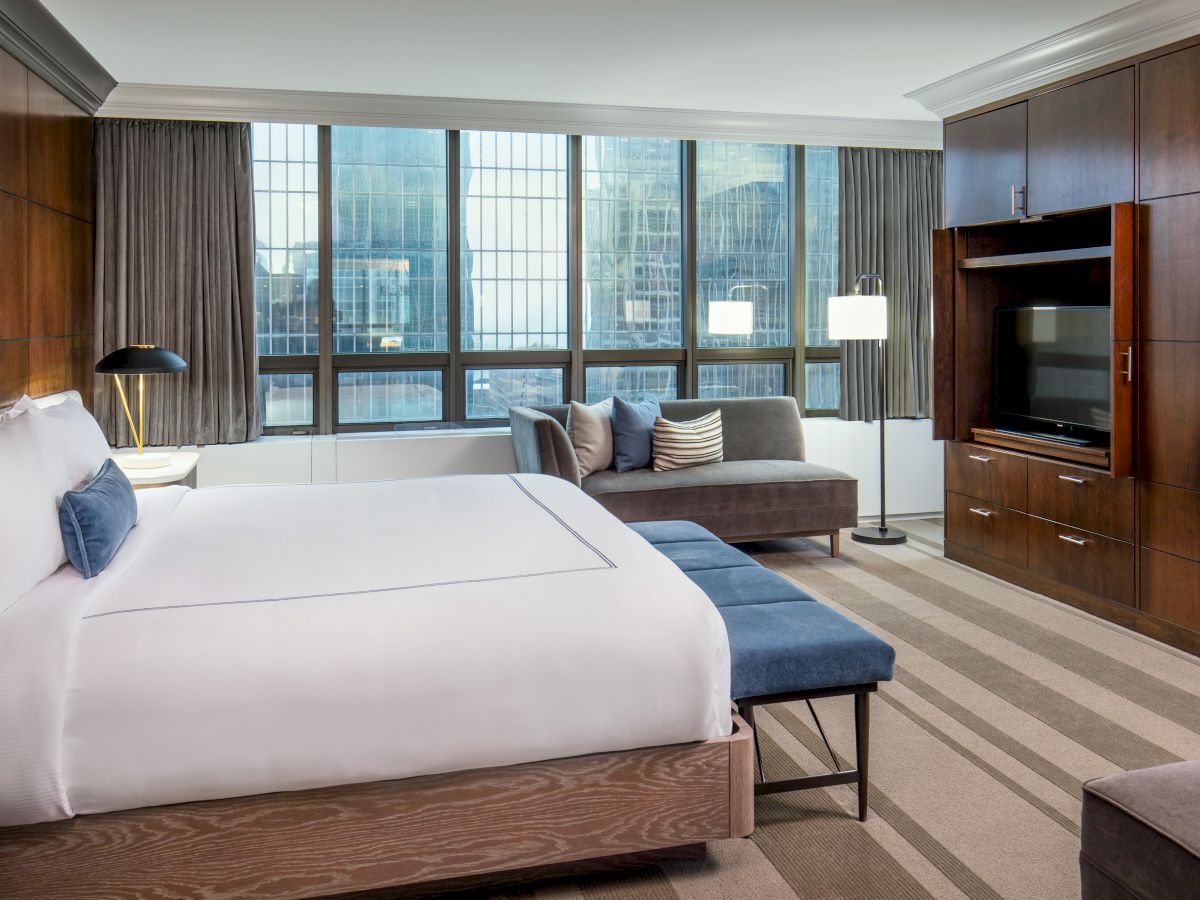 A modern hotel room featuring a large bed, a sofa, a TV, a large window with city views, and a wooden decor theme, ending the sentence.